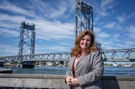 Woman with brown hair wearing gray suit st和s in front of Memorial Bridge in Portsmouth.